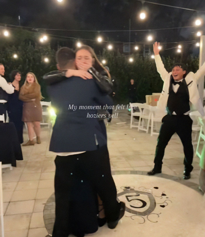 Couple embracing during a proposal at a wedding, with guests reacting in the background.