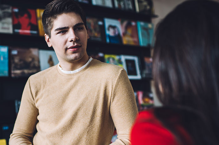19-year-old in bookstore, wearing a beige sweater, talking to a person in red, with shelves of books in the background.