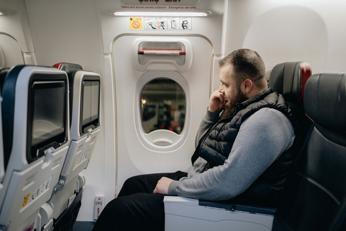 Man sitting by airplane window, looking thoughtful, wearing a black vest, related to "woman made 3 men move" scenario.