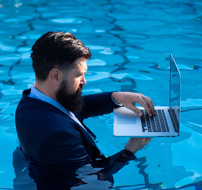 Man in a suit using a laptop while standing waist-deep in a pool, symbolizing work commitment even on vacation.