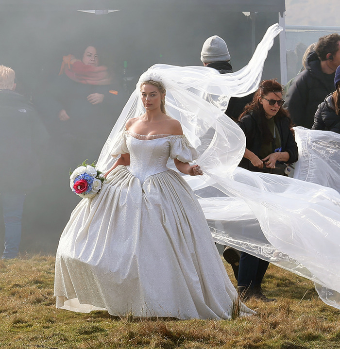 Actress in elaborate Wuthering Heights costume holding flowers, with crew in the background on a misty set.