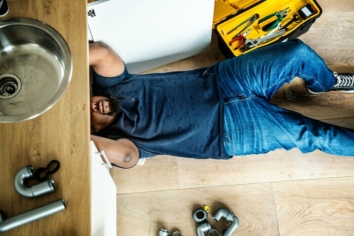 Man fixing pipes under sink, tools scattered, wearing jeans and a dark shirt, illustrating a humorous realization about partner’s skills.