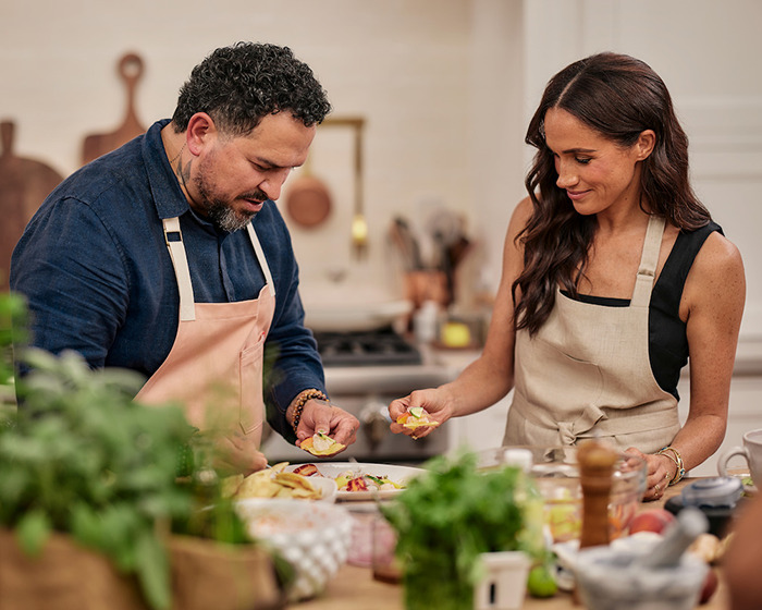 Meghan Markle with guest cooking together in a kitchen, wearing aprons and surrounded by ingredients. Meghan Markle with guest cooking together in a kitchen, wearing aprons and surrounded by ingredients.