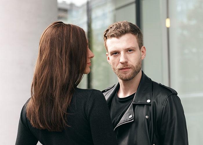 Man in a leather jacket gazing ahead, while a woman in a black top stands nearby, representing questions about men.