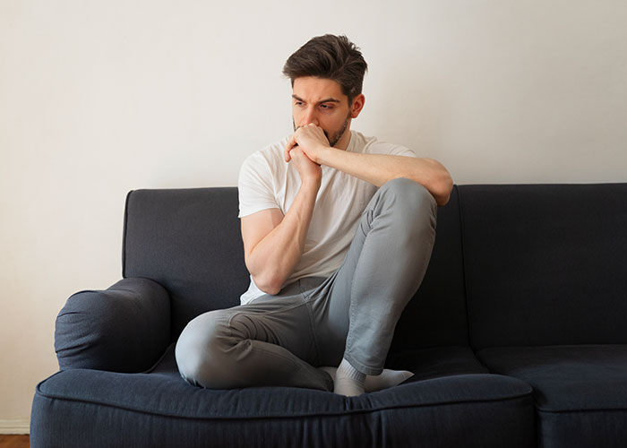 Young man in deep thought sitting on a couch in a casual home setting, reflecting on questions about men.