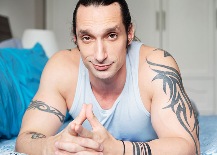Man with tattoos lying on a bed, wearing a light blue tank top, and looking at the camera.