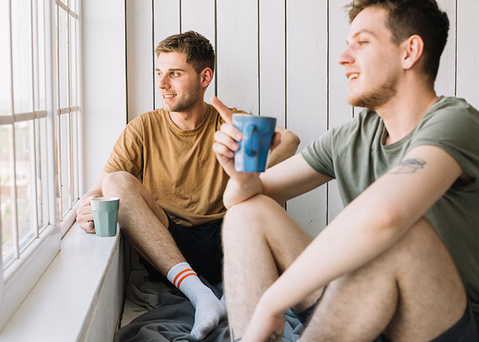 Two men sitting by a window, holding mugs, and chatting casually.