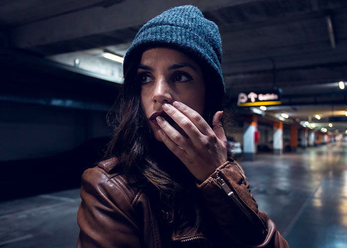 Woman in a parking garage, wearing a blue beanie and brown jacket, pondering questions about men.