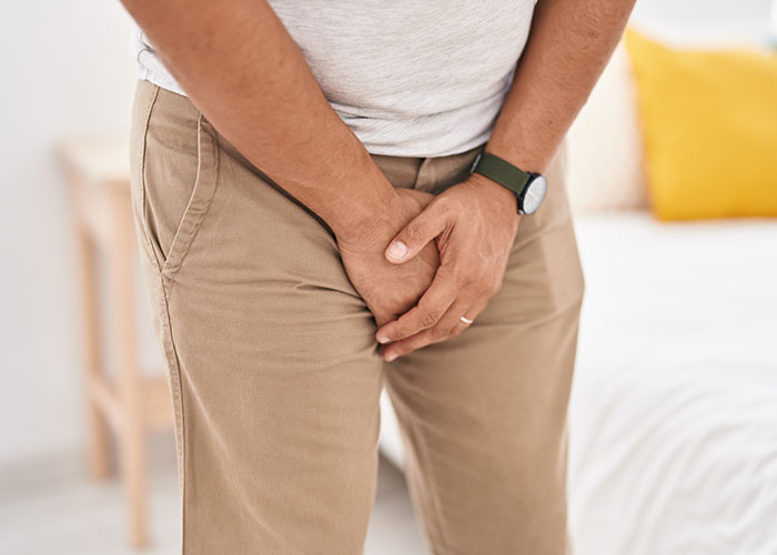 Man in beige pants holding his groin area, standing in a bedroom, addressing common questions about men.