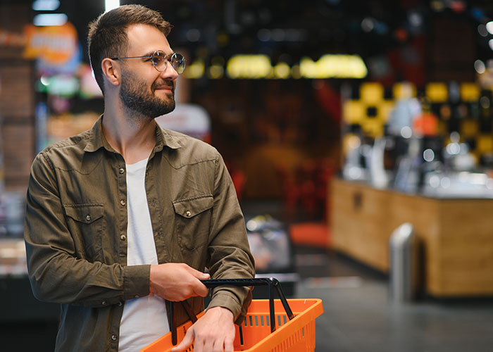 Man with a beard and glasses shopping in a grocery store, holding an orange basket, reflecting on questions about men.