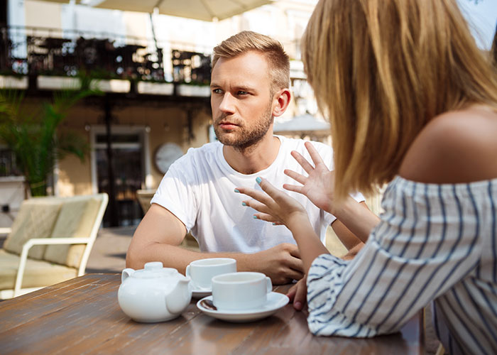 Man listening to woman at outdoor café, discussing questions about men, with cups and teapot on table.