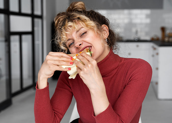 A woman in a red sweater enjoying a wrap in a modern kitchen, smiling with curiosity about men.