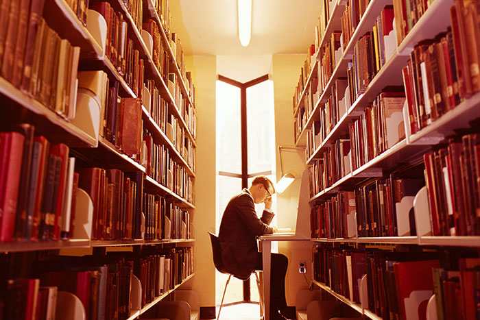 Man sitting at a library desk, surrounded by books, focusing on reading under warm light.