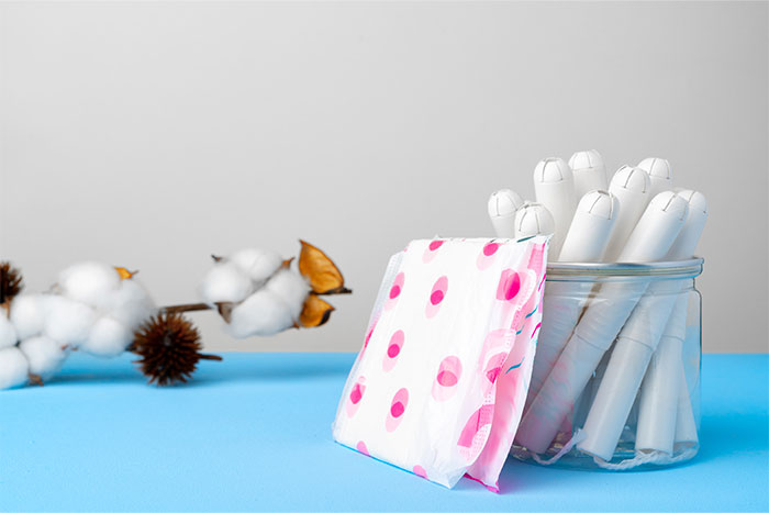 Toallas higiénicas y tampones en un frasco sobre fondo azul, con ramas de algodón decorativas.