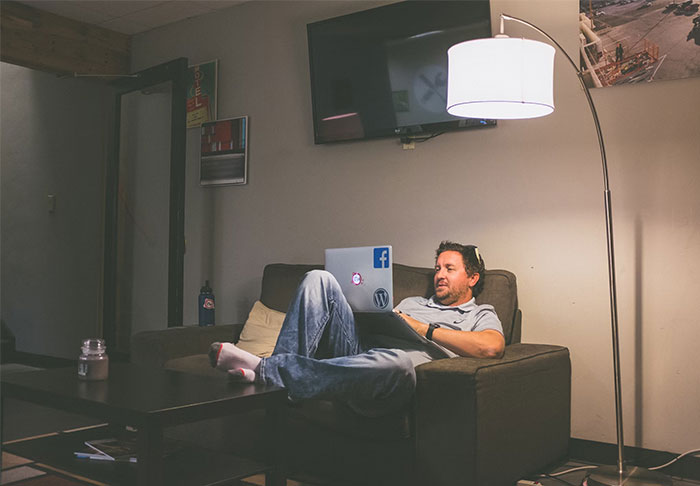 Man lounging on a sofa with a laptop, under a lamp in a cozy room, relaxed atmosphere.