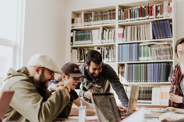 Men laughing around a table in a library, engaging in a discussion, possibly involving questions they find embarrassing.