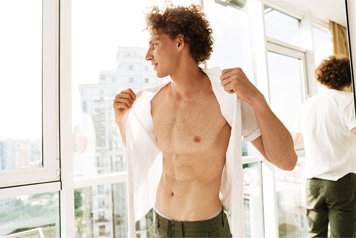 Man standing indoors, unbuttoning shirt, looking away, with cityscape view.