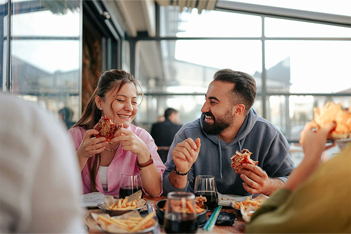 Man and woman enjoying burgers and fries, sharing a laugh at a casual dining table, with drinks in front of them.