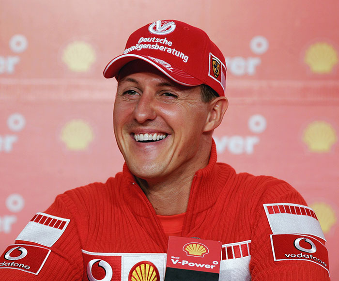 F1 legend in red cap and jacket, smiling at a press event, with Shell and Vodafone branding in the background.