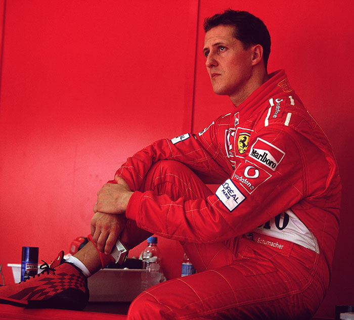 F1 Legend Michael Schumacher in Ferrari race suit seated, deep in thought.
