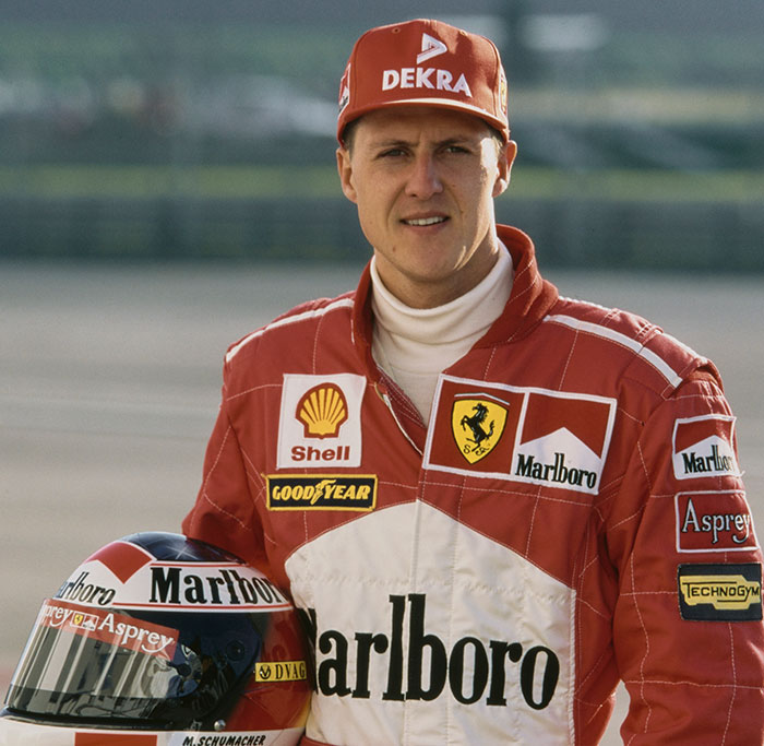 Man in a red racing suit and cap holding a helmet, representing F1 legend Michael Schumacher.