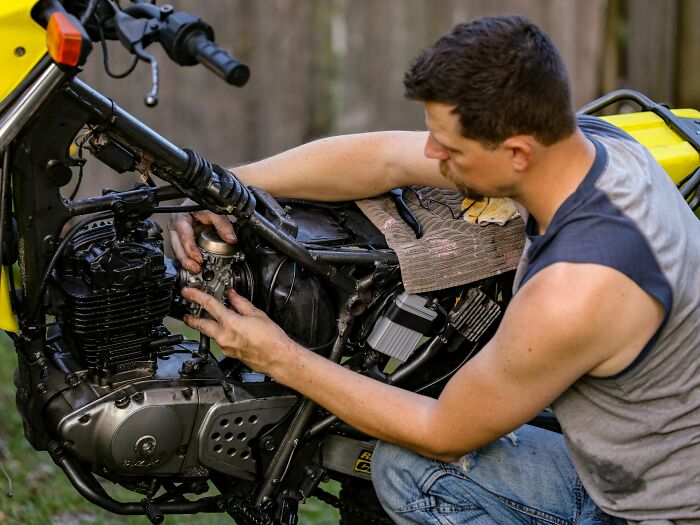 A man working on a motorcycle engine, focusing on the engine parts, outdoors in casual attire.