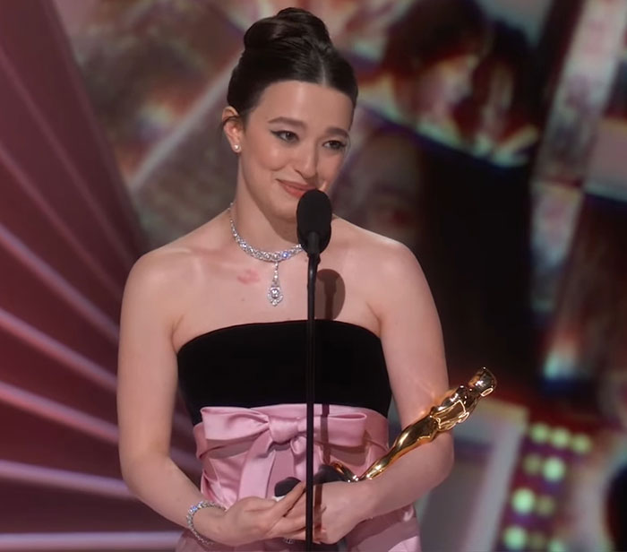 Young woman holding an Oscar statue, wearing a pink and black gown, standing at a microphone on stage.