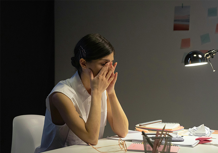 A woman sits at a desk, looking stressed while planning wedding invitations under a desk lamp.