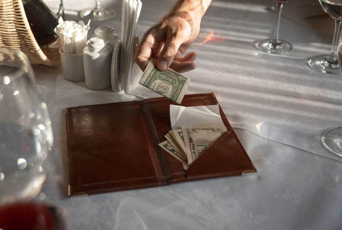 Hand placing cash in a brown bill holder on a restaurant table, highlighting wallet absence. Hand placing cash in a brown bill holder on a restaurant table, highlighting wallet absence.