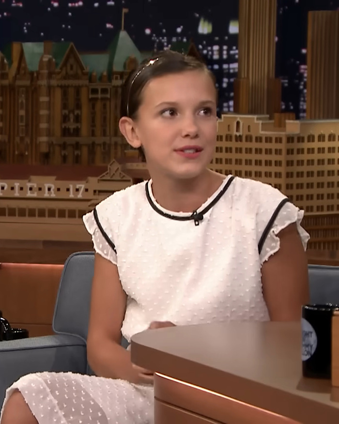 Young Millie Bobby Brown in a white dress on a talk show. Young Millie Bobby Brown in a white dress on a talk show.