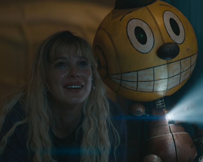 Blonde woman with an excited expression next to a large yellow robot, emitting light.