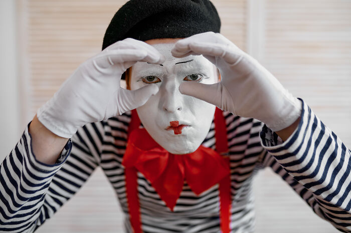 Mime performer with white face paint and black beret enacting a gesture, wearing a striped shirt and red bowtie.