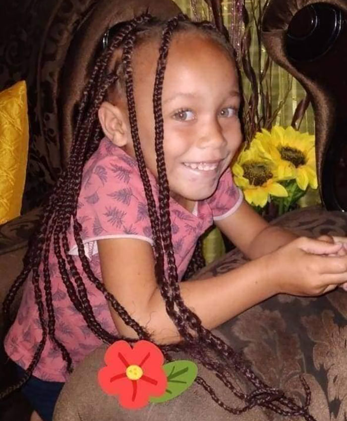Young girl with light eyes and braids, smiling on a couch with sunflowers in the background.