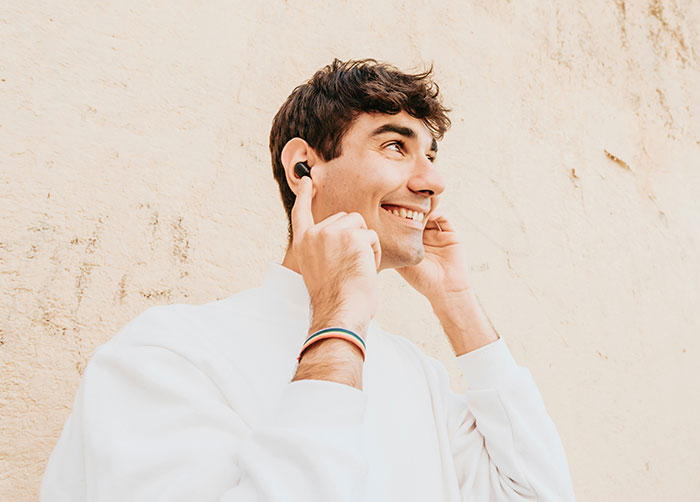 Man smiling, using wireless earbuds against a beige wall, showcasing modern conveniences.