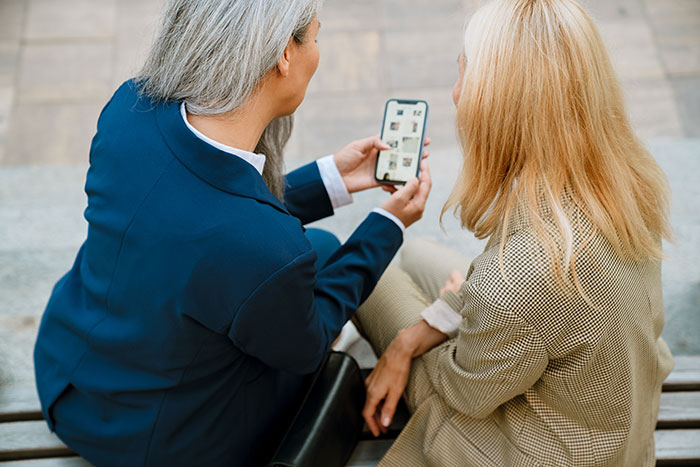 Two women discussing modern conveniences while looking at a smartphone outdoors.