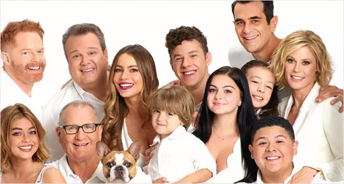 A group of people in white attire posing together, with Ed O'Neill and a dog in the front.