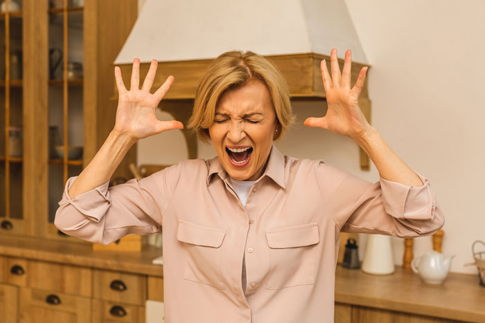 A woman in a kitchen showing frustration with her hands raised and eyes closed.