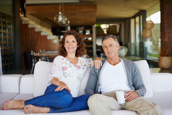Couple sitting on a white couch in a modern living room, lady in floral blouse and jeans, man holding a coffee cup.