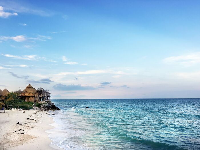Stunning beach with clear blue waters, white sand, and coastal huts under a serene sky.