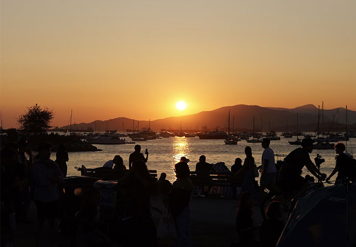 Silhouetted group by water at sunset, boats and mountains, capturing a memorable coincidence moment.