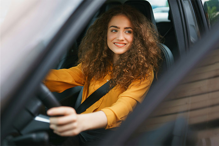 Woman with curly hair, wearing a yellow shirt, smiling while driving a car, embodying the spirit of bridesmaids' experiences.