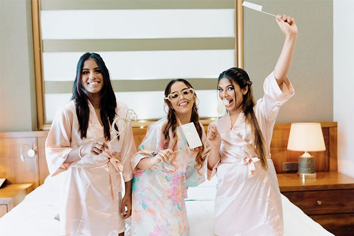 Bridesmaids in robes smiling, holding props, and posing playfully in a cozy bedroom setting.