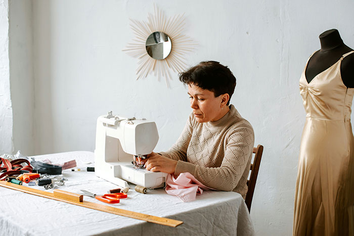 A woman sewing a pink dress, with sewing tools on the table, illustrating a bridesmaid's task.