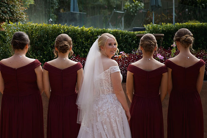 Bride smiling with bridesmaids, all in burgundy dresses, standing outdoors before the ceremony.