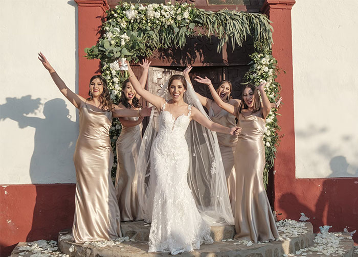 Bride with bridesmaids in champagne dresses under a floral arch, celebrating joyfully.