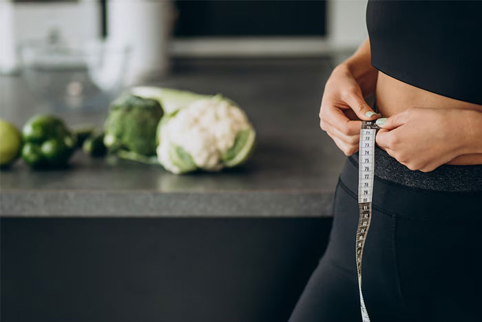 Woman measuring waist with tape measure, vegetables on kitchen counter, illustrating bridesmaids' ridiculous requests for fitness.
