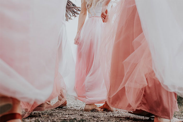 Women in pastel pink dresses walk on gravel, showcasing bridesmaids.