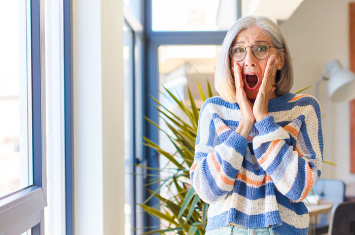 Surprised woman in colorful sweater with hands on face, standing by a window. Surprised woman in colorful sweater with hands on face, standing by a window.