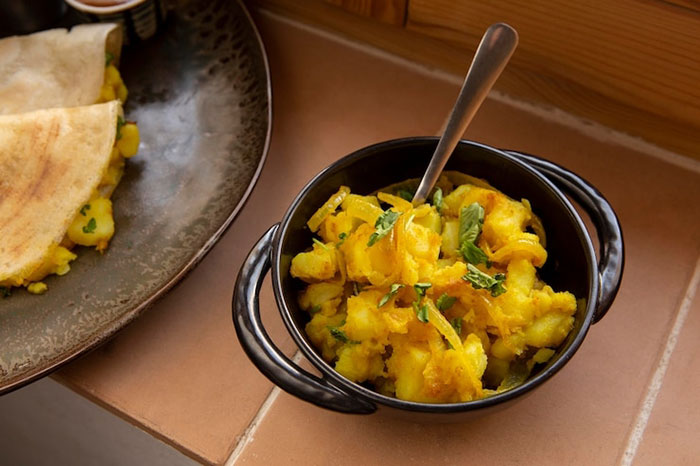 Homemade Indian food in a black bowl with a spoon, featuring a savory potato dish, on a tiled surface.