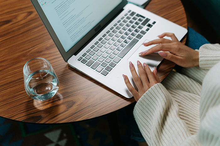 Woman typing on a laptop at a round table, glass of water nearby, focusing on a story about shattered dreams.